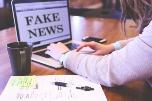 Una detective sentada a la mesa con su portátil, toma notas mientas en su pantalla aparece el título "Fake News". Imagen de memyselfaneye en Pixabay