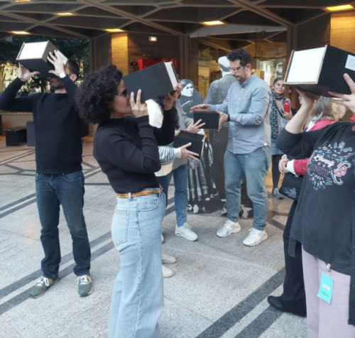 Grupo de personas en un espacio interior sosteniendo cajas negras frente a sus ojos, como si observaran a través de ellas. La escena sugiere una actividad participativa o artística, con varios asistentes interactuando y un ambiente de taller o exposición.