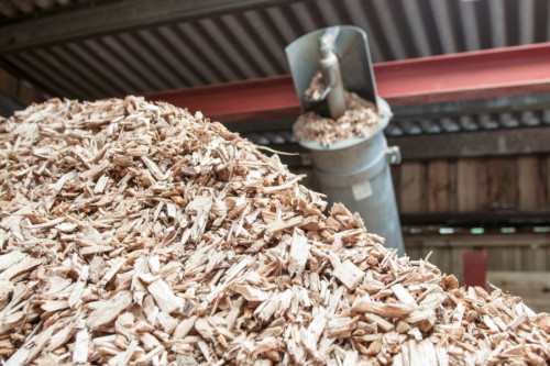 Aquí tienes la traducción del texto de la imagen:  **Texto original:**  > "Close-up of a large stack of wood chips in a hopper, for use in a heating boiler, with an automated auger above the stack." >  >   **Traducción al español:**  > «Primer plano de un gran montón de astillas de madera en una tolva, para su uso en una caldera de calefacción, con un sinfín automatizado sobre el montón.»