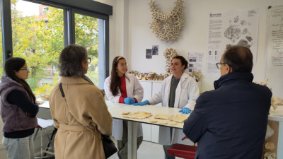 En un laboratorio luminoso, dos personas con bata blanca explican unas muestras de biomateriales sobre la mesa a un pequeño grupo de visitantes que las rodean y escuchan de pie.